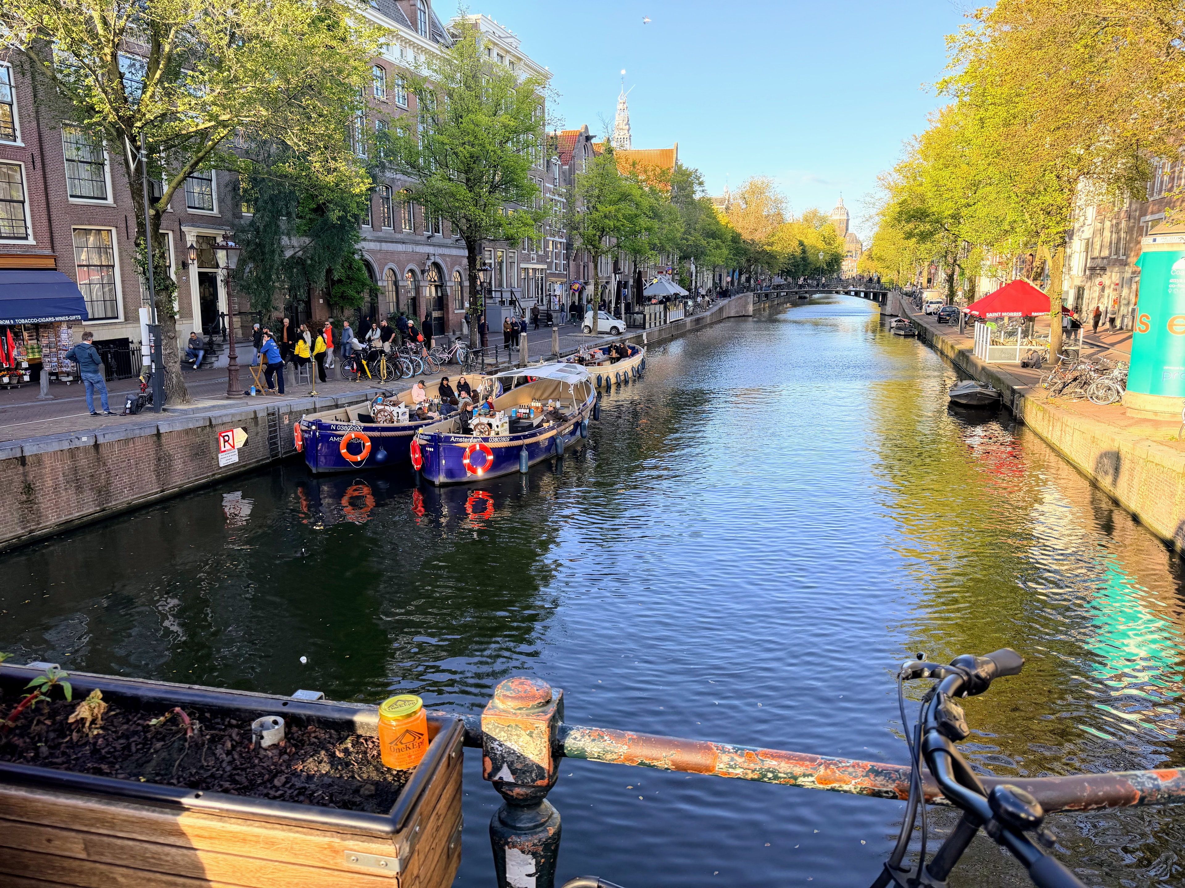 OneKEpp honey jar on a bridge railing overlooking an Amsterdam canal with tour boats and the Westerkerk spire